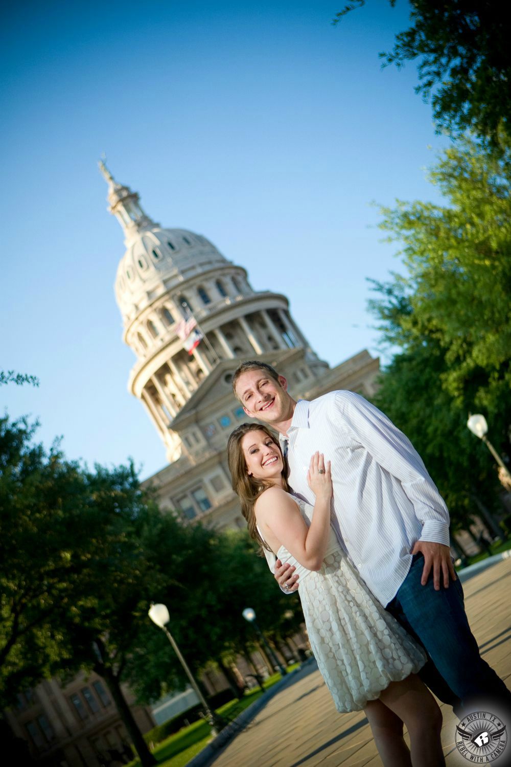 confident, happy girl with dark hair in light shirt and large beaded necklace stands and holds in front of a stoic guy with dark hair and a dark shirt in a stairwell at the Texas Capital in this romantic black and white engagement picture in Austin.
